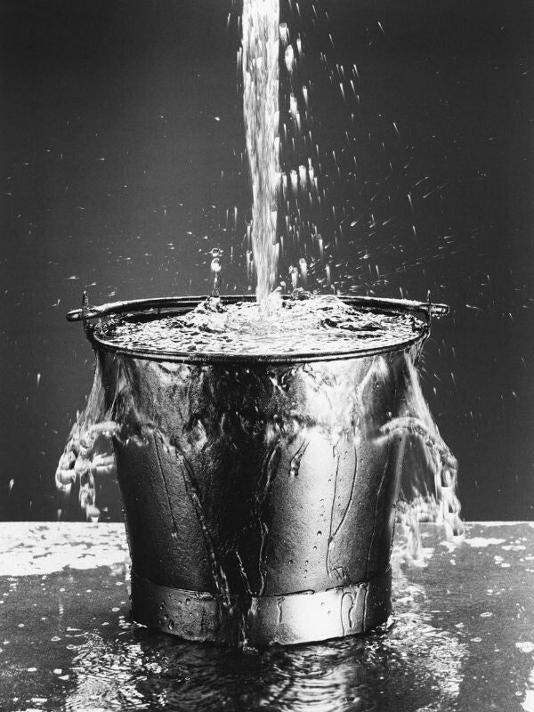 Emmer die bijna overloopt doordat uit verschillende bronnen waterdruppels vallen als metafoor voor de optelsom van werk, privé en leefomstandigheden die kunnen bijdragen aan verzuim.