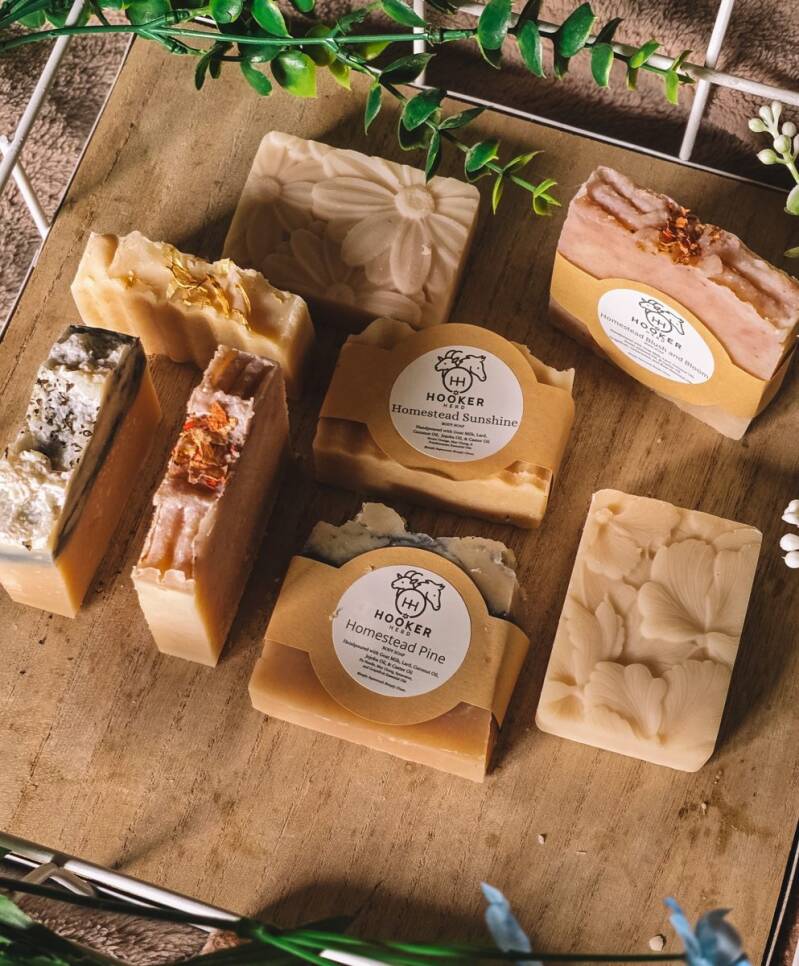 Hooker Herd goat milk soap bars with labels, including Homestead Sunshine, on a wooden tray with greenery