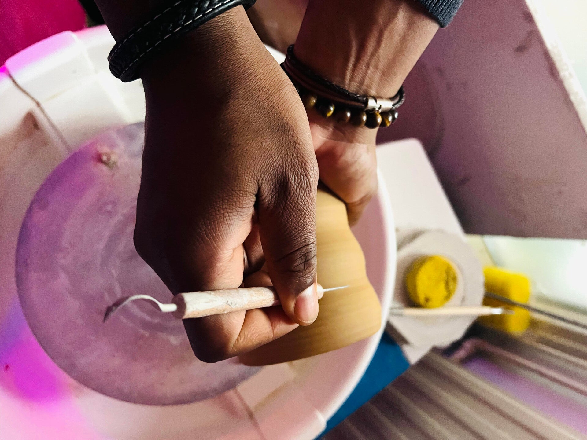 Een paar handen die met klei gereedschap een mok aan het bewerken zijn met op de achtergrond een draaischijf