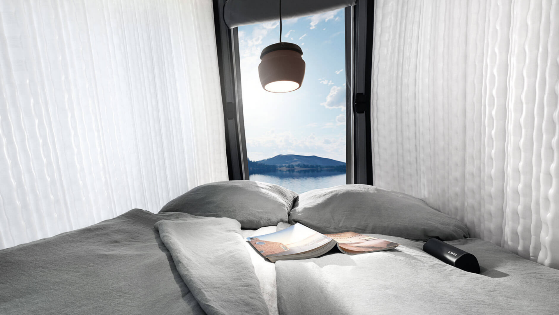 Lit confortable avec vue sur un lac et des montagnes depuis une chambre lumineuse avec livre ouvert, ambiance cosy et nature.