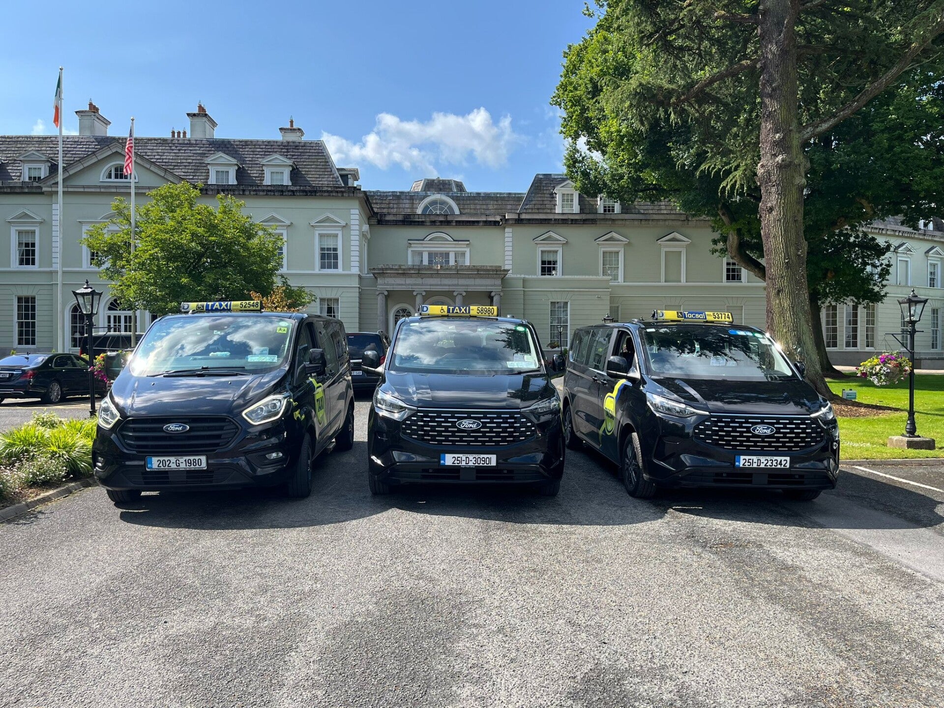 3 new black taxi minivans parked side by side, facing forwards in front of a hotel. Green tree to the right. Blue sky. forwards