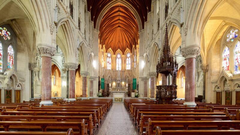 Interior of St. Colman’s Cathedral in Cobh, Ireland, showing its high vaulted wooden ceiling, ornate stone columns with red marble shafts, detailed Gothic arches, carved wooden pulpit, and colorful stained glass windows illuminating the long central aisle