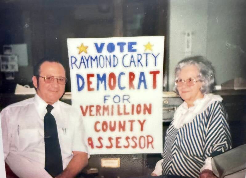 Man with white shirt and glasses with a sign and an grey-haired woman.