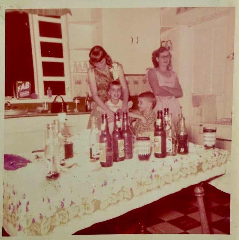 Two women in a kitchen with two children, the table is covered with empty beer bottles.