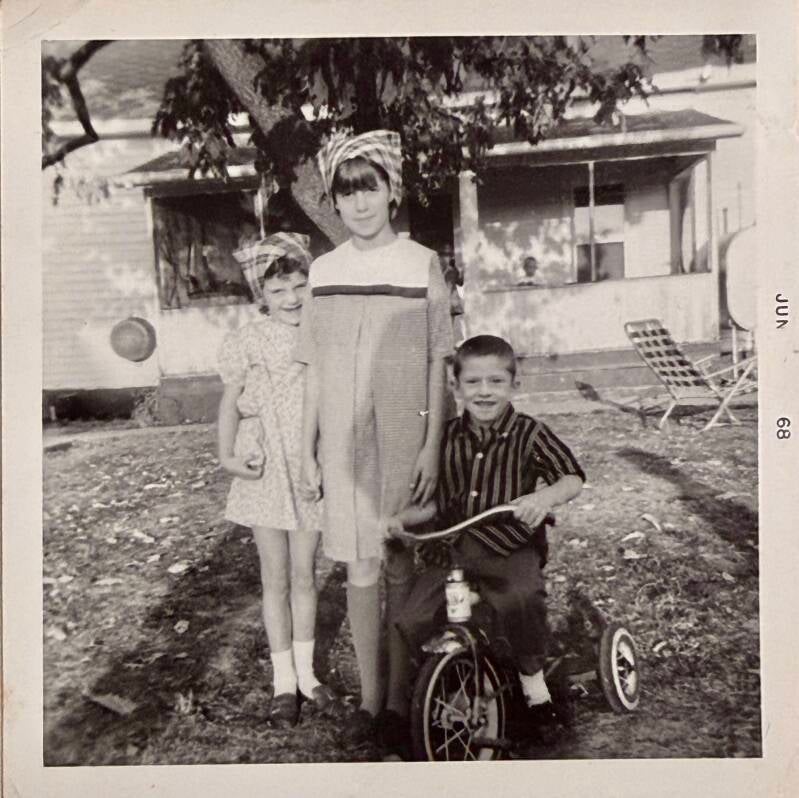 Three siblings posing. The sisters are holding hands and the boy is on a tricycle. 