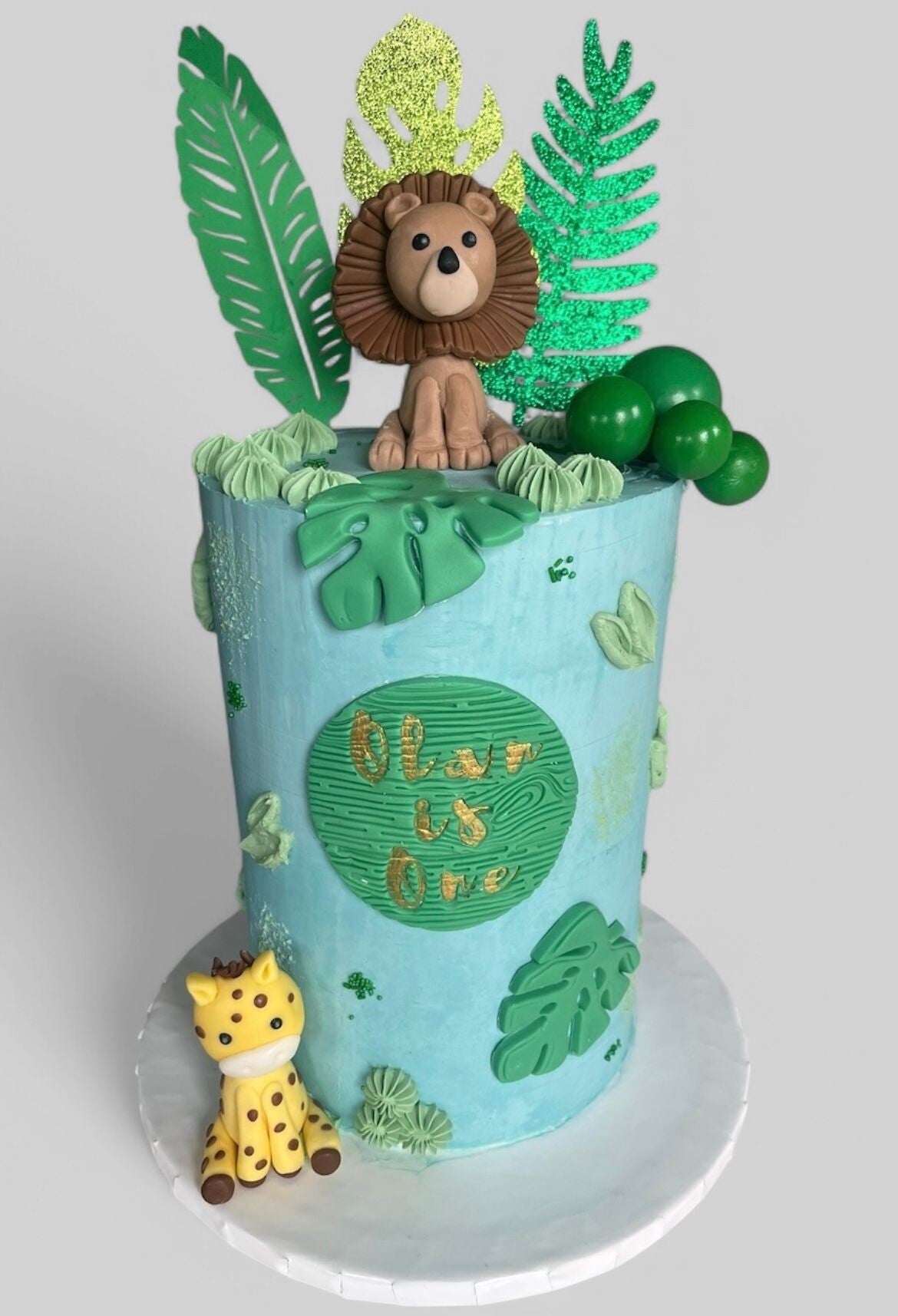 Jungle themed 1st birthday cake, tall with a lion and giraffe fondant figures and decorated blue and greeen. 