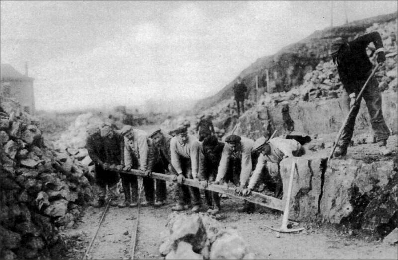 Op de foto wrikken 9 mannen om een mergelblok los te krijgen uit de mergelgroeve van Kroongroeven. Een stukje smalspoor van de kalkbranderij is zichtbaar. Op de achtergrond is een stukje van het vroegere seinhuis van Schin op Geul te zien. Links liggen me