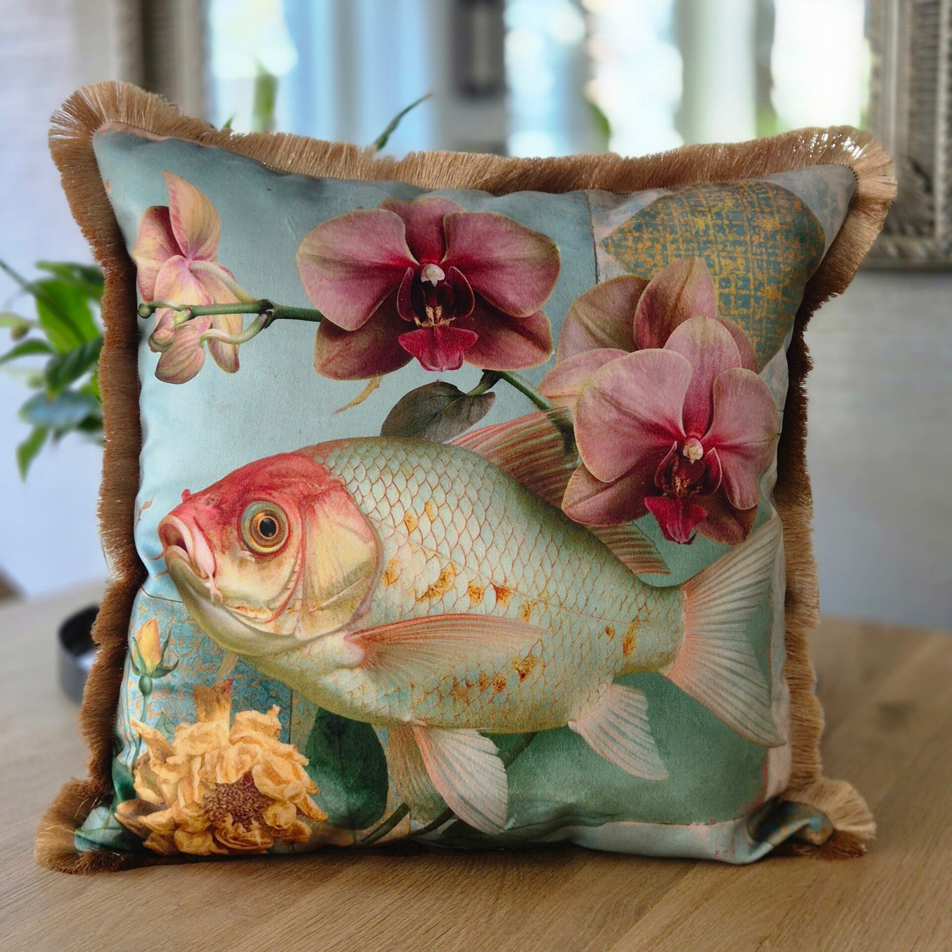 Sierkussen met een gedetailleerde print van een grote, zilver-rode vis en donkerroze orchideeen op een lichtblauwe achtergrond. Het kussen is afgewerkt met een goudkleurige franje.