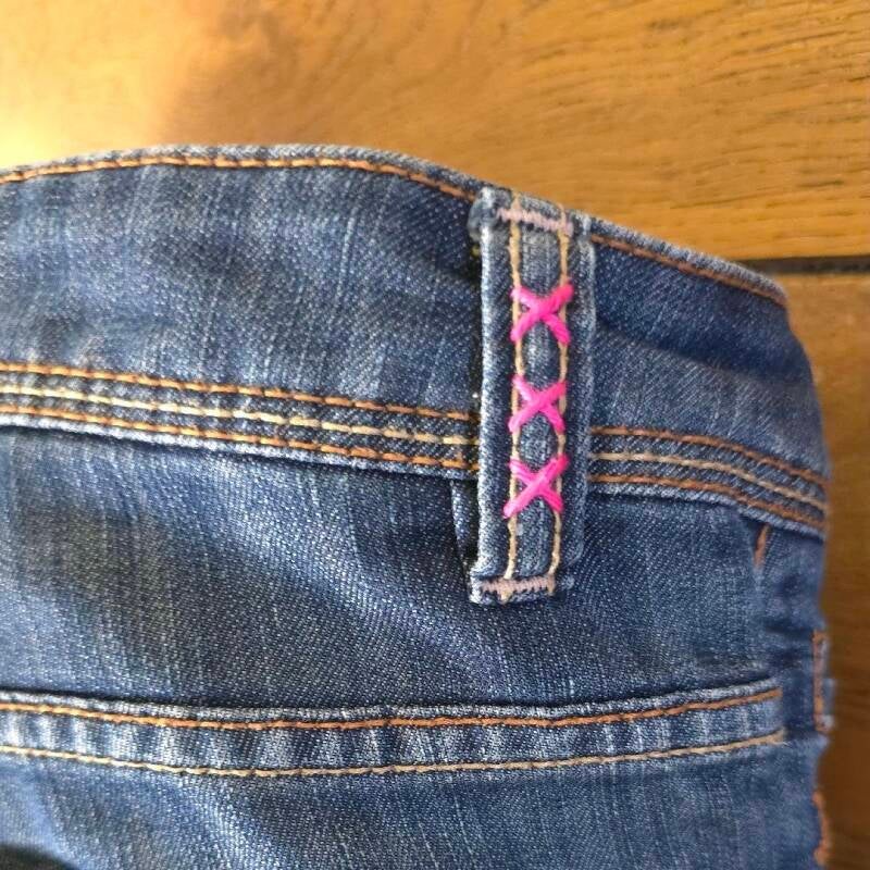 Close-up of embroidered belt loops with three pink crosses – Hand-stitched pink crosses on denim belt loops, inspired by Amsterdam’s coat of arms.