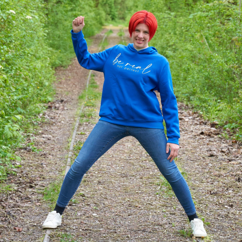 Stephanie Breschniak steht in einer kraftvollen Pose. Auf dem blauen Pullover ist der Aufdruck "be real not perfect". Mein Lebensmotto