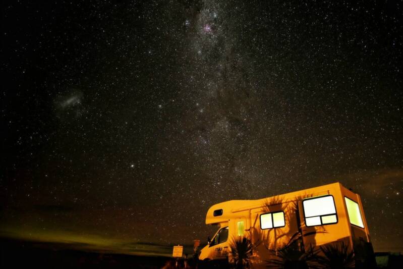 Campervan with windows aglow from internal lights. Above the Van the milky way shines brightly and with a clarity that is hard to find in our world showing the freedom of air pollution that acompanies travel throughout new zealand 