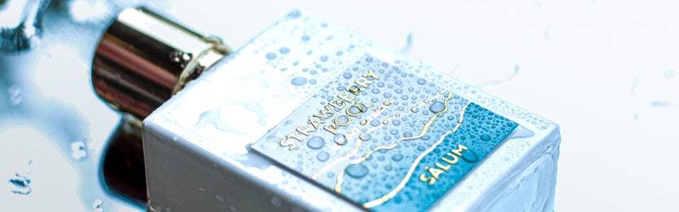 Close-up of Strawberry Pool by Salum Parfums — a white perfume bottle with gold cap, covered in water droplets, lying on a reflective wet surface. The blue and gold label evokes a poolside vibe, matching the summery, aquatic identity of the fragrance.
