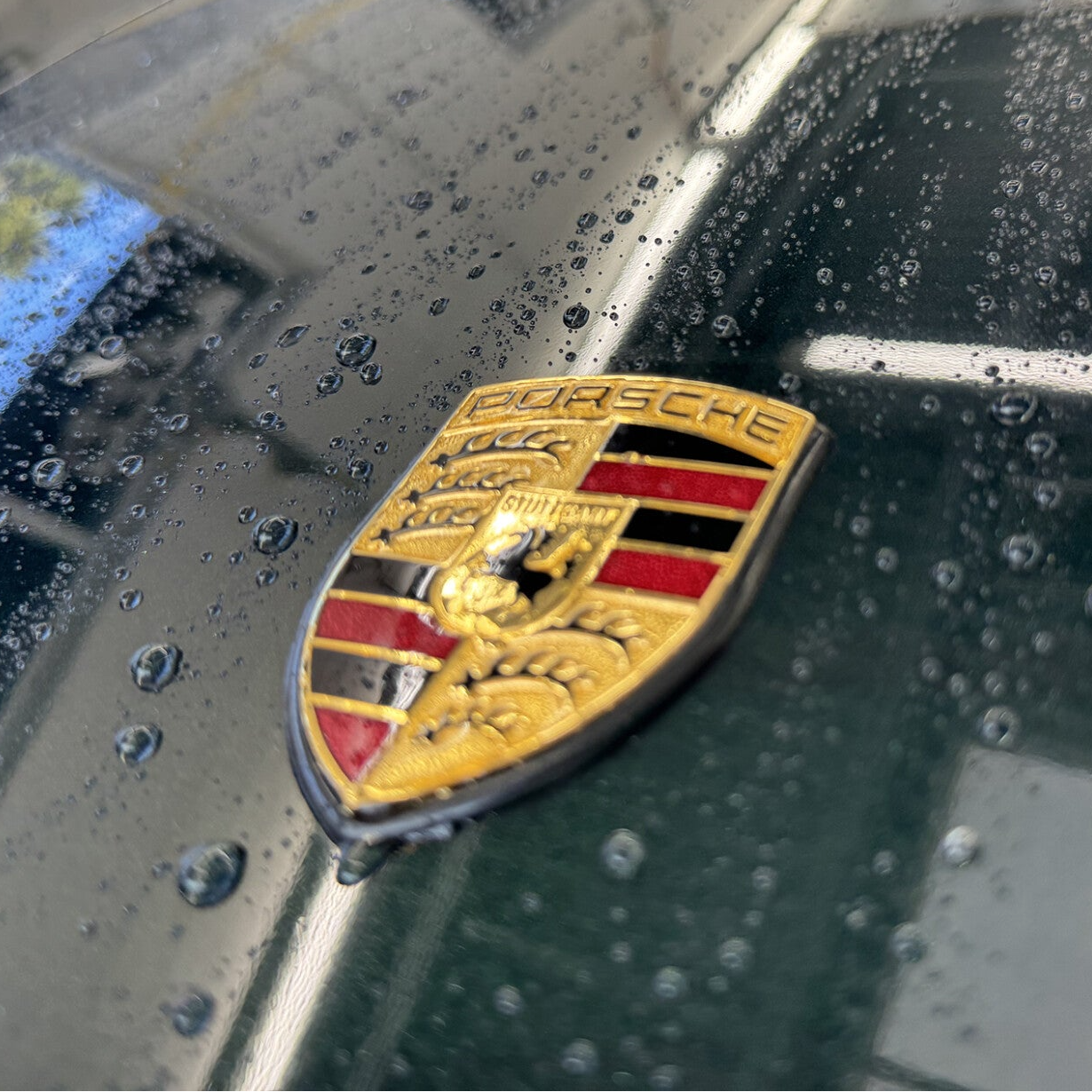 a close up image of a porsche logo on a car with water dropplets around the logo 