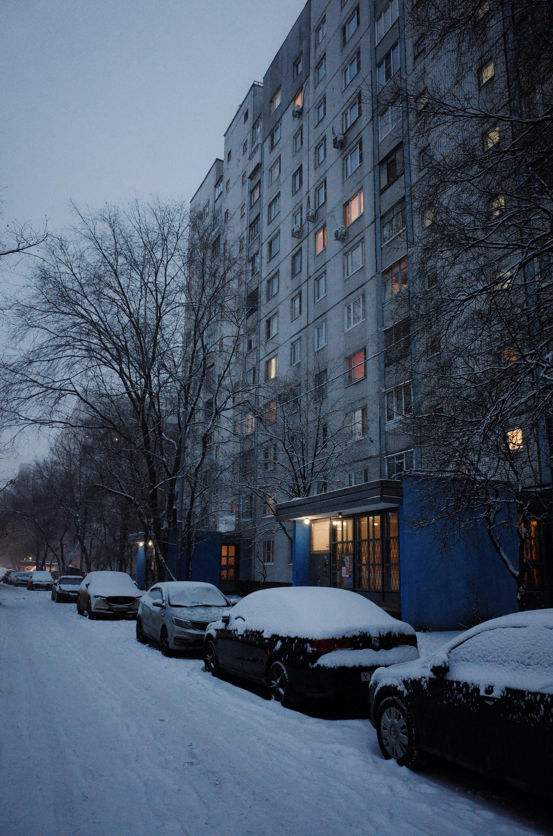 Cars parked on the side of the road, in front of a blue building, with snow covering the road and the top of the cars 