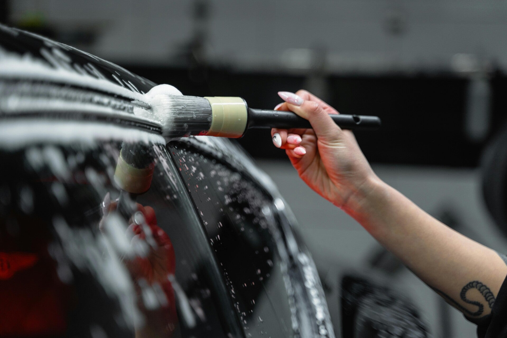 a close up of a womens hand holding a brush with soap bubbles on it cleaning the top of a car window 