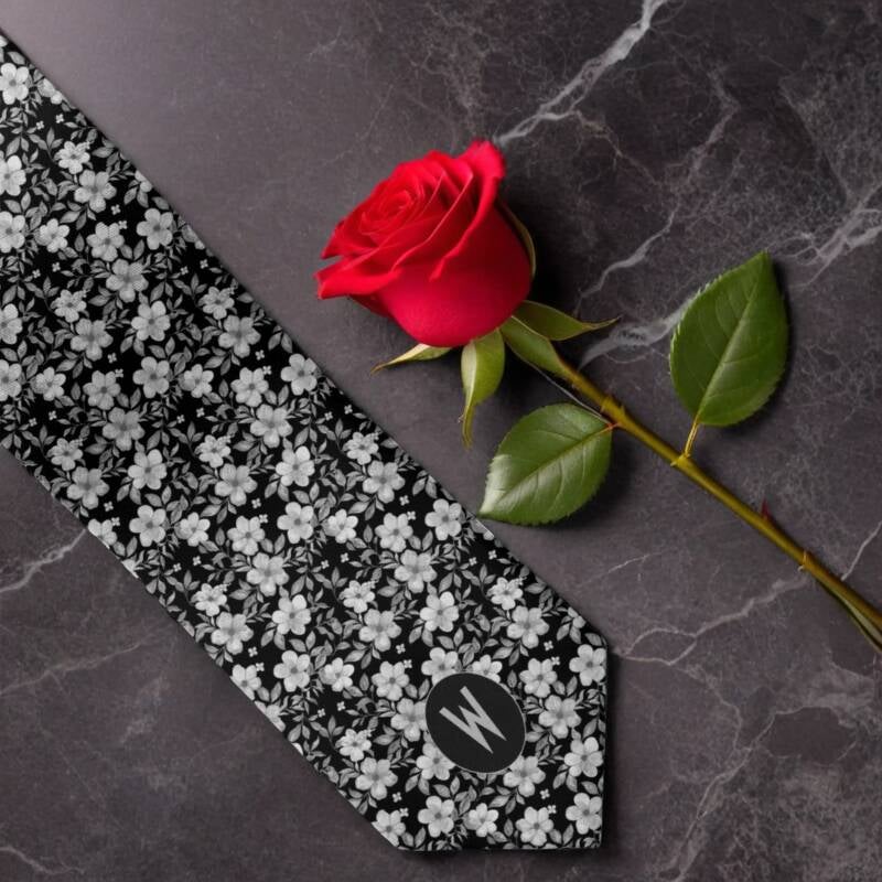 Black-and-white floral wedding groomsmen's necktie with monogram lying on a marble table top next to a red rose.