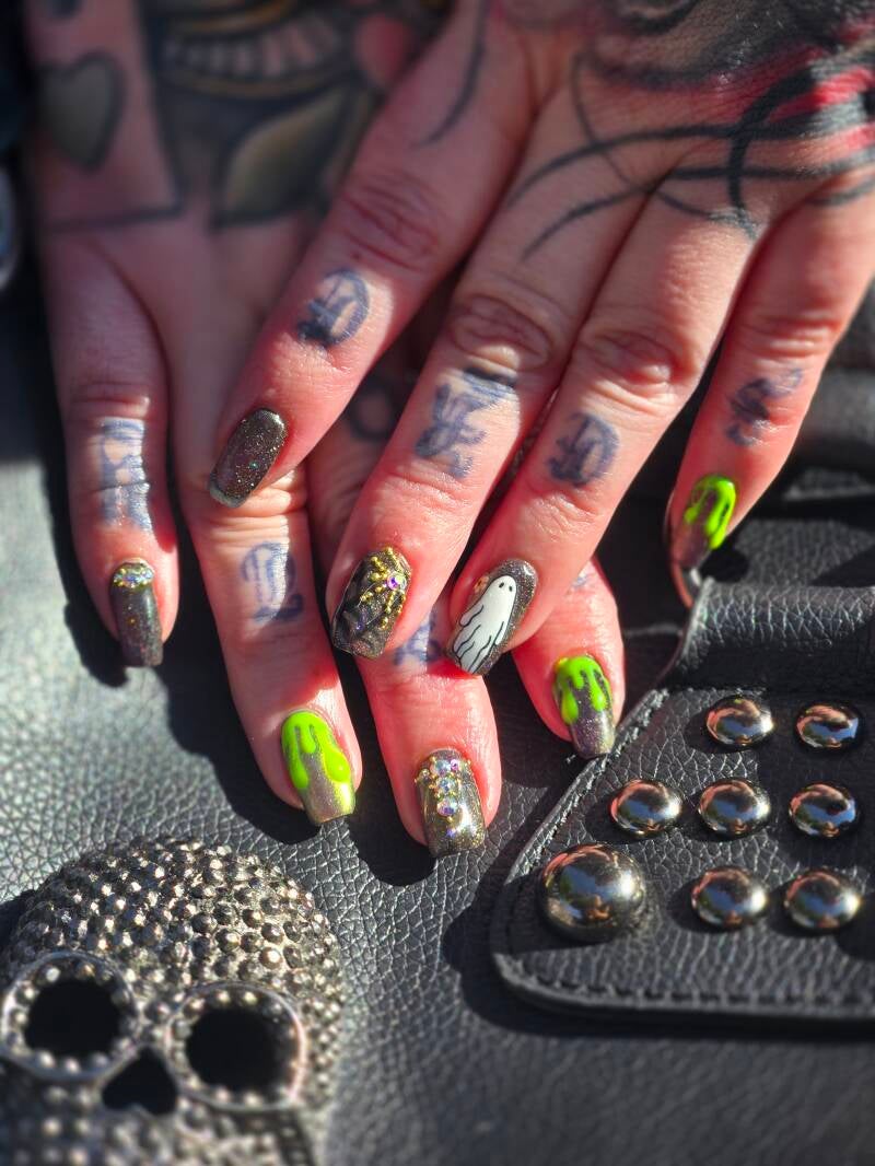 Close up of tattooed woman's hands atop a black leather skull purse the vibe of the nails is Halloween with a dark base green neon blood drips ghosties and chrome with spiders made with gold balls and lots of rhinestones 