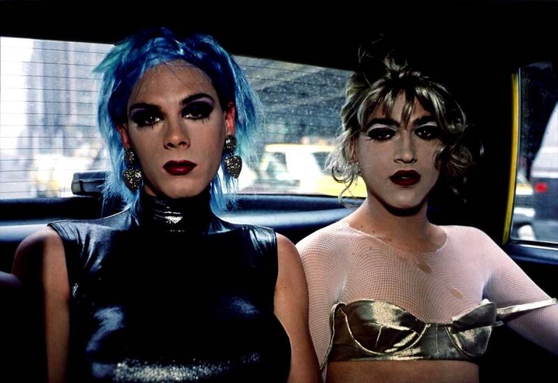 A blue haired drag queen in a sparkly dress and a blond draw queen in a gold bra and net top