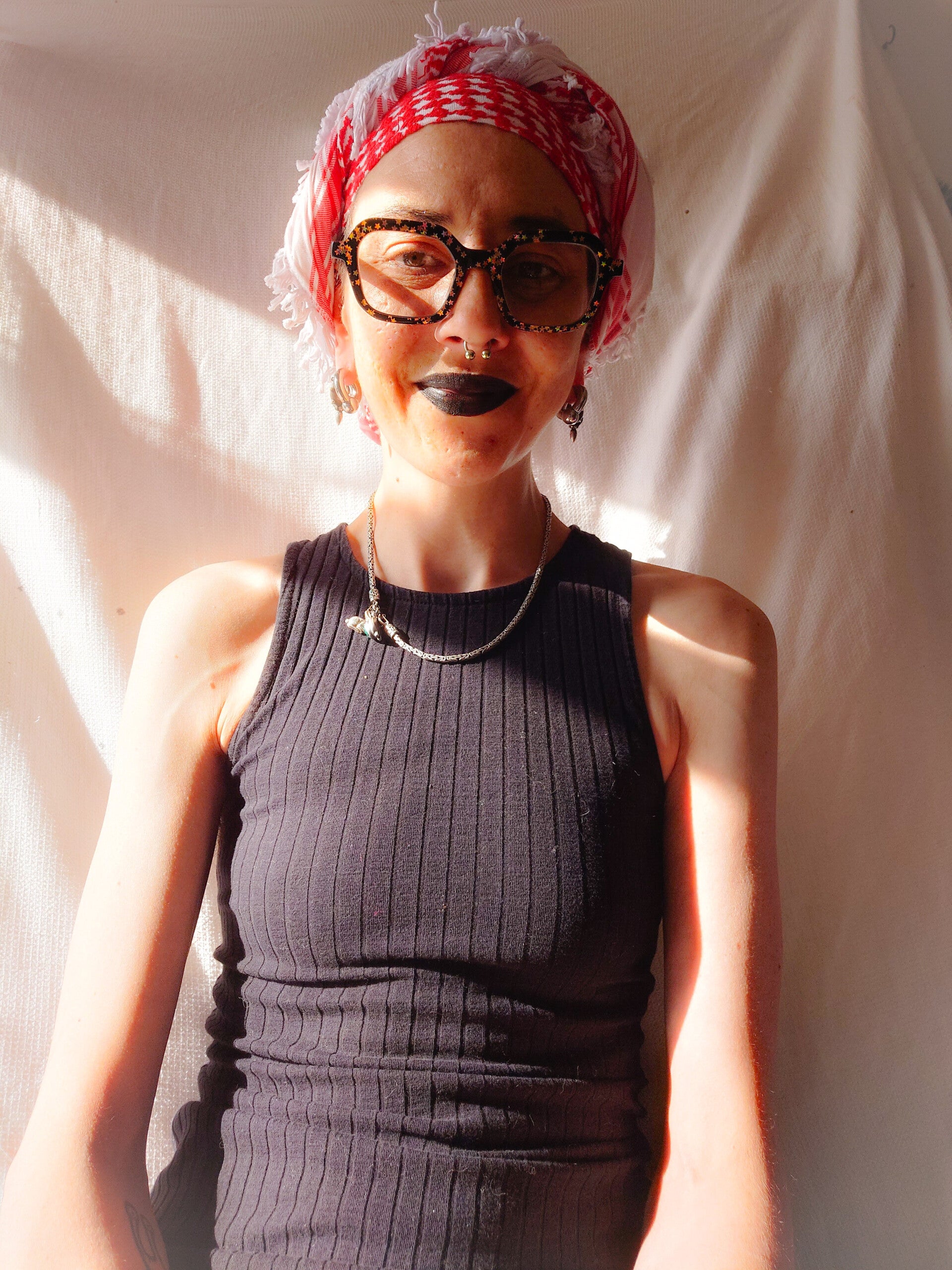 A white woman with a red and white headscarf, black square Elton John glasses with star motif and a black top. She has a septum piercing and black lipstick on.
