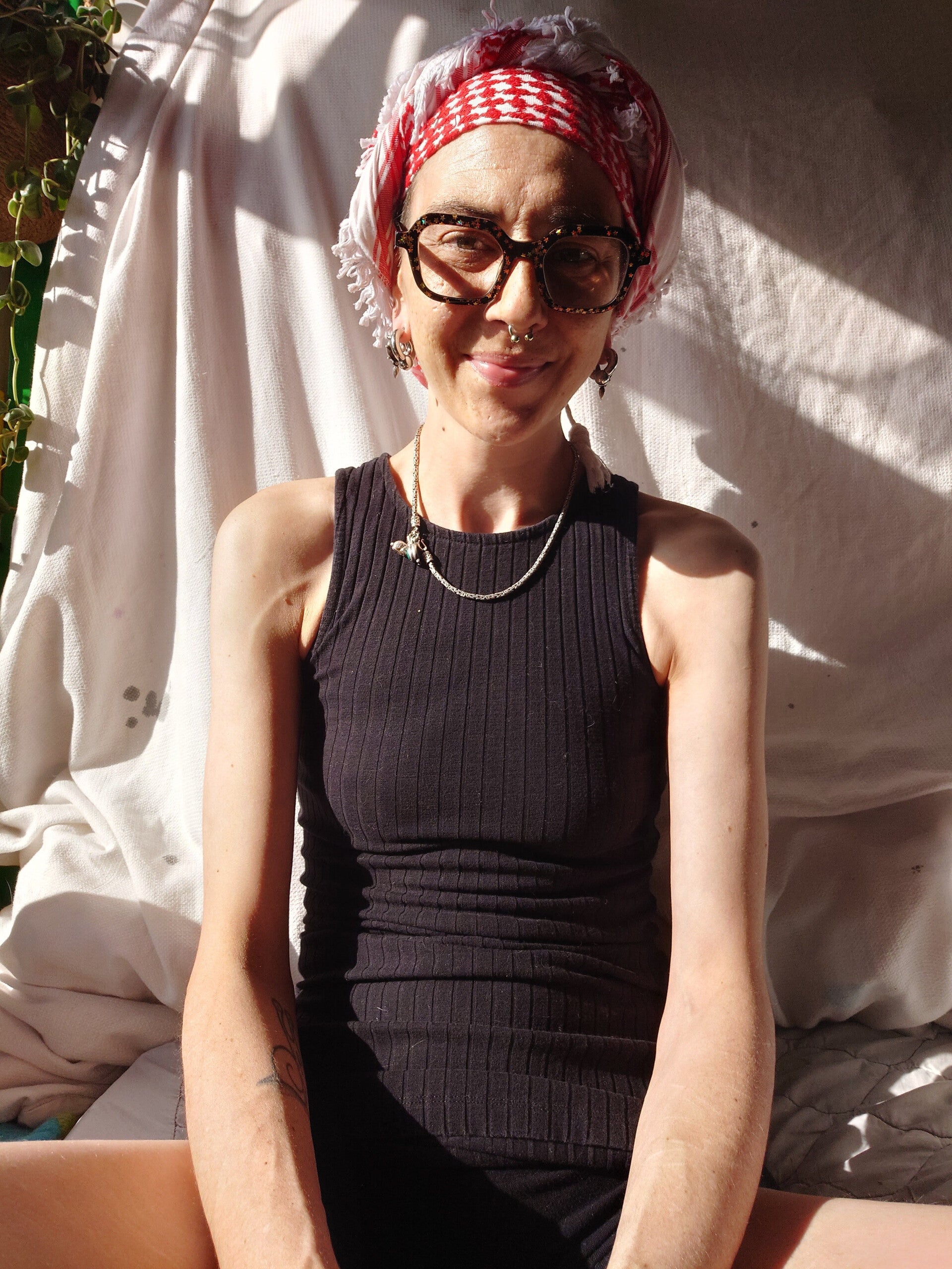 A woman with a red and white headscarf sits in front of a white screen. Behing them is a plant, they wear black square specs and a black top 