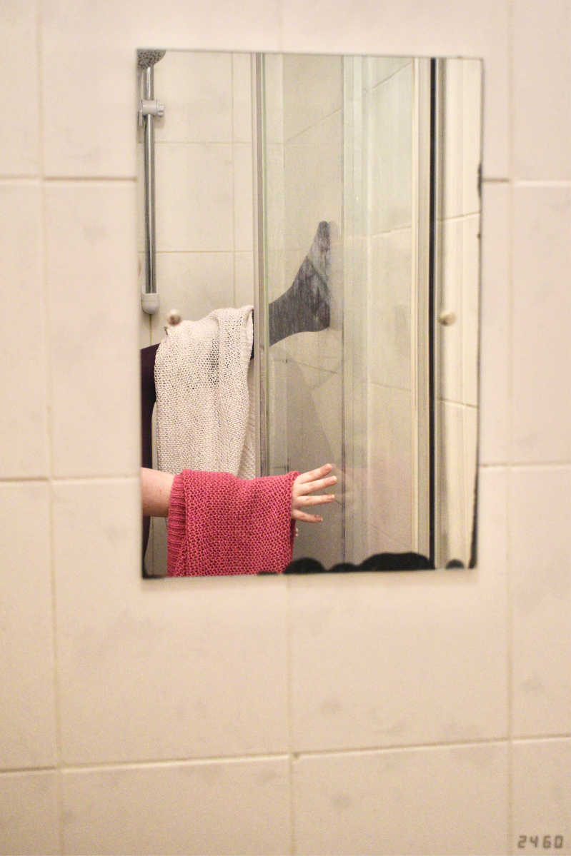Bathroom mirror with leg in purple tights, pale-skinned arm, and white and pink jumpers