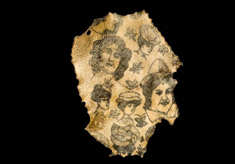 Pezzo di pelle tatuata umana antica con ritratti e disegni, conservata come reperto storico, raffigurante volti femminili e motivi floreali in bianco e nero su sfondo marrone.