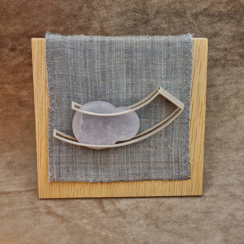 This brooch is made of sterling silver, blue alabaster and a nickel silver brooch pin displayed on a stand out of oak and linen.