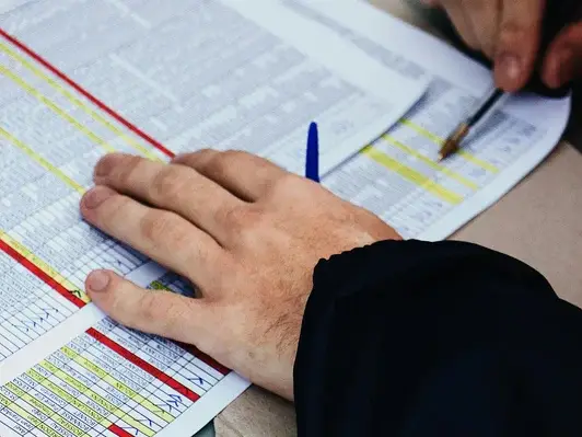 A close-up shot of a person's hands reviewing a horse racing form. One hand rests on the page while the other holds a pen, pointing at specific entries highlighted in yellow.
