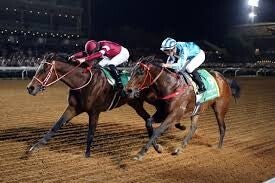 Two jockeys in a tight race at night on a dirt track. One jockey wears maroon and white silks, while the other wears light blue and white. Their horses are captured mid-stride under stadium lights with a crowd visible in the background.