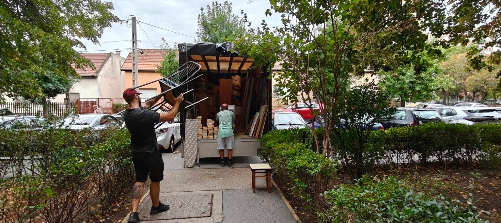 Ponyvás teherautó hátulról, nyitott ponyvával, rakodás közben, egy férfi a platón lévő bútorokat rendezi és egy másik két összerakott széket visz a platóhoz