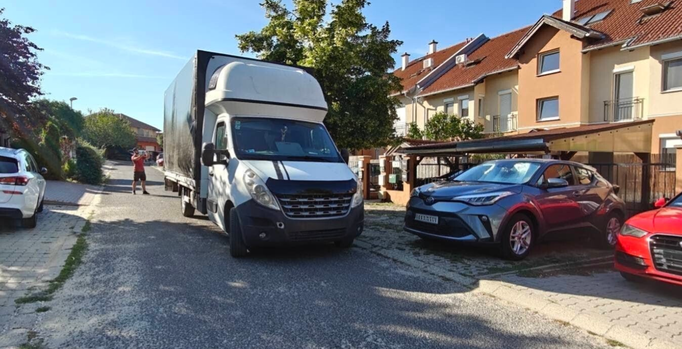 Biztosfuvar ponyvás teherautó egy kertvárosi utcán parkol autók mellett a házak előtt napsütésben, egy kollega segít a pontos beállásban