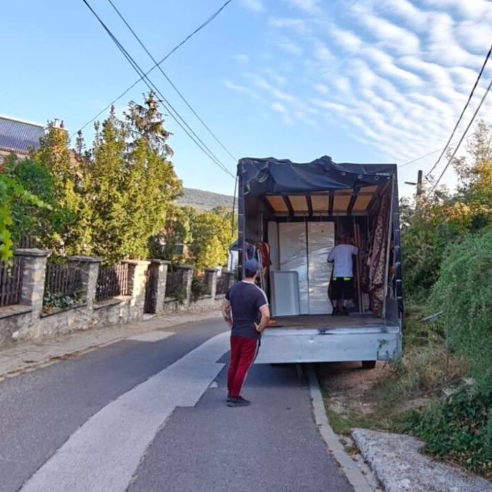 Két férfi bútorokat rakodik a Biztosfuvar teherautó platójára őszi, napsütéses időben egy kertes övezetben