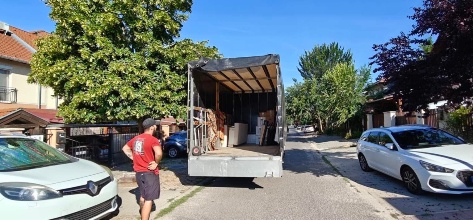 Biztosfuvar ponyvás teherautó hátulról, nyitott pinyvával rakodás közben, az autó mögött egy kolléga várakozik