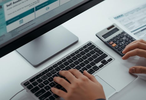 Close-up of hands typing on keyboard with HMRC Self-Assessment portal on screen, documents and calculator aside, titled ‘Self-Assessment Tax Return Explained’”