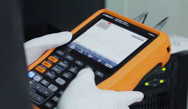 A Siglent SHS1000X handheld oscilloscope being used in the field with gloved hands