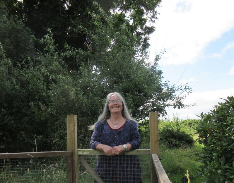 An older  woman leaning on a wooden gate in a wildish bit of garden.