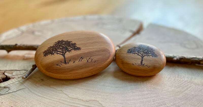 Zwei Holz-Handschmeichler mit Lebensbaum-Motiv auf einer Holzscheibe – beide zeigen eine unterschiedlich entwickelte Version des Motivs.