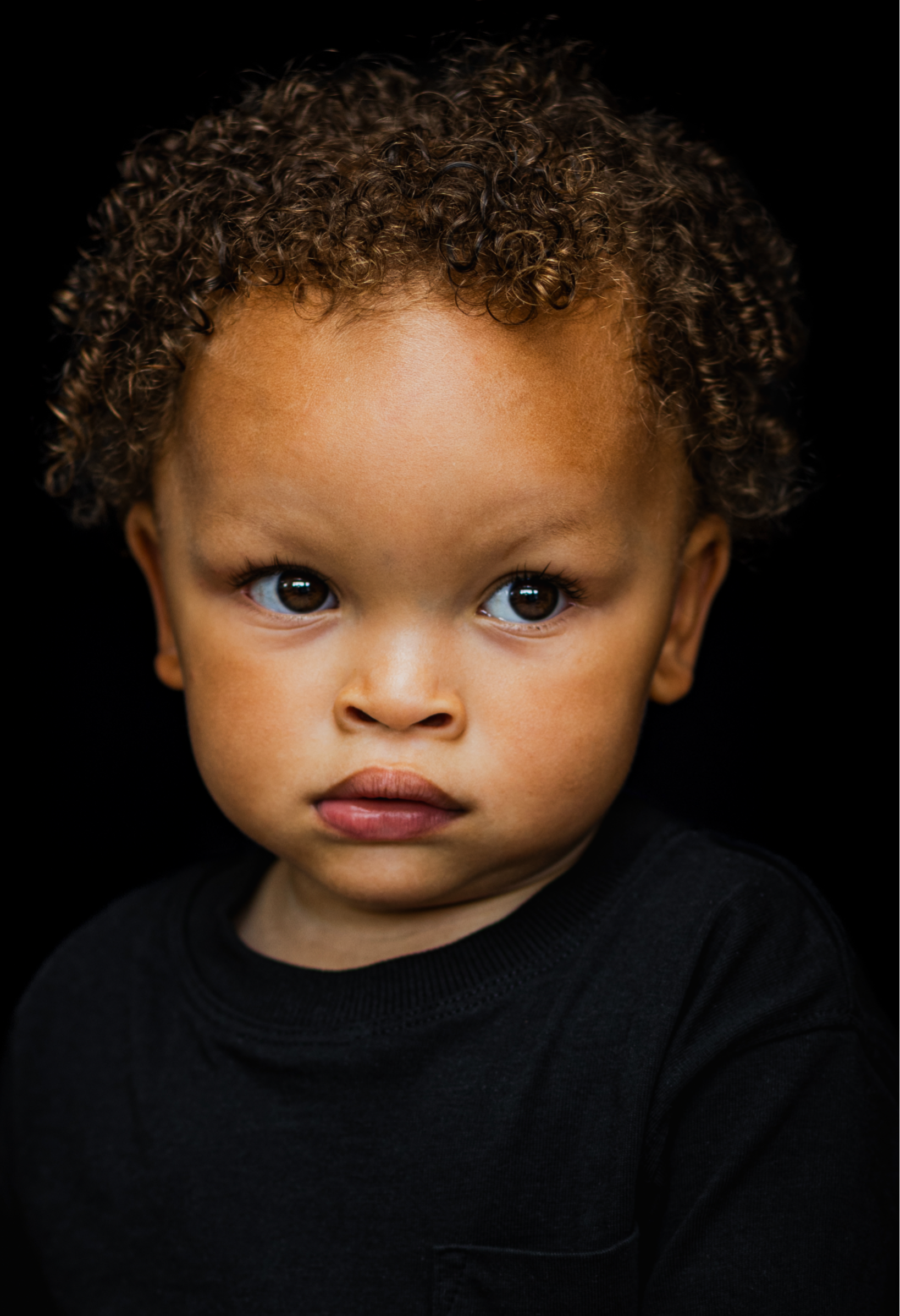 limlachend jongetje met donkere krullen op een portretfoto met zwarte achtergrond – kinderfotografie door Alyssa Dijkstra