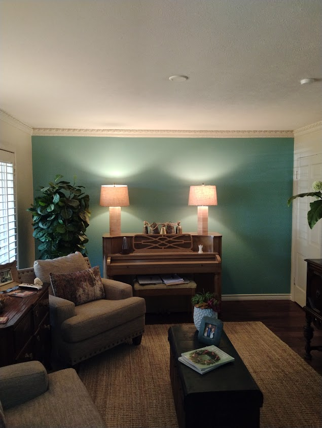 A cozy living room with a dark green accent wall, a piano, warm lighting from two lamps, and a seating area featuring a sofa and a coffee table with decor elements