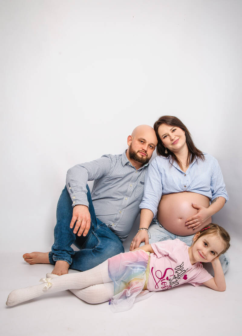 Przyszli rodzice z córeczką podczas studyjnej sesji ciążowej w Grudziądzu – mama w rozpiętej błękitnej koszuli eksponuje brzuszek, a dziewczynka leży na pierwszym planie