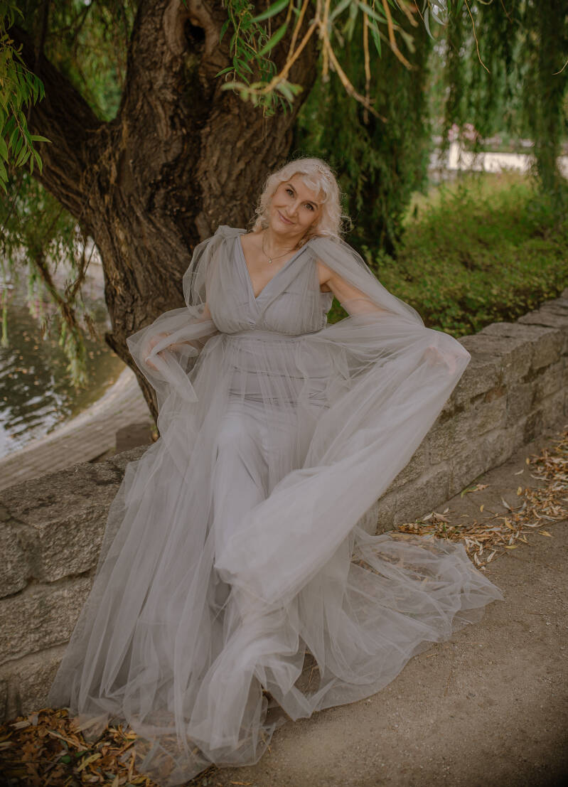  – elegancka sesja kobieca przy fontannie i stawie blisko Chełmna oraz Świecia. Dynamiczny portret dojrzałej modelki w szarej stylizacji glamour na tle zielonych gałęzi wierzby.