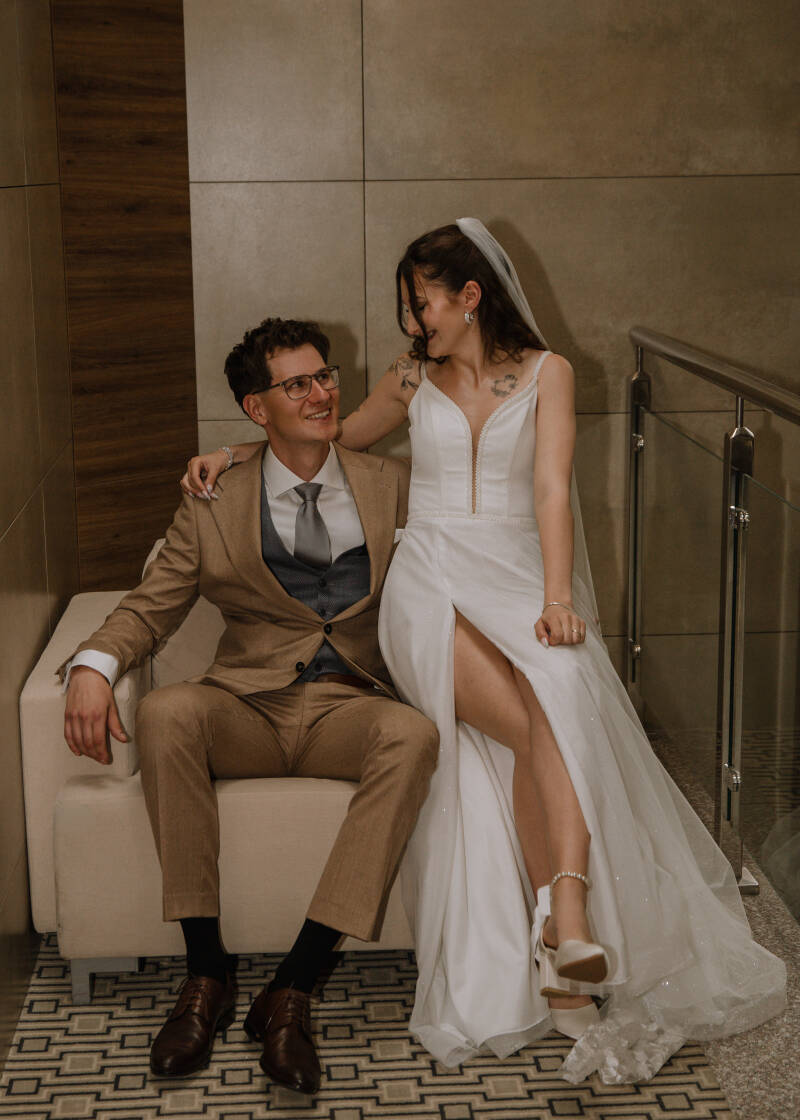 Fotografia ślubna Grudziądz – nowoczesna mini-sesja ślubna w Hotelu Rudnik. Pan młody w brązowym garniturze siedzi w jasnym fotelu, a panna młoda w sukni z głębokim rozcięciem siedzi na oparciu, eksponując nogę i buty ślubne.