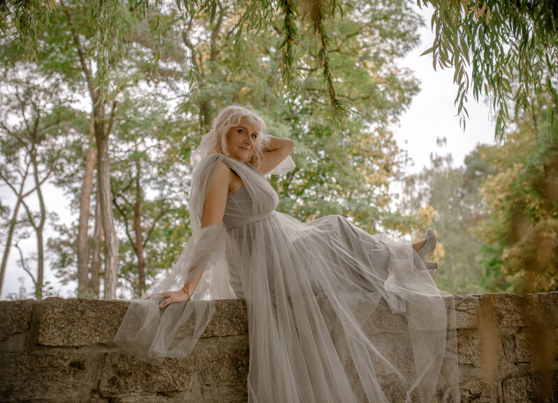 Fotograf Grudziądz i okolice – plenerowa sesja urodzinowa dla kobiet blisko Bydgoszczy i Torunia. Elegancki portret dojrzałej modelki w zwiewnej szarej stylizacji glamour, siedzącej na kamiennej barierce nad parkowym stawem.