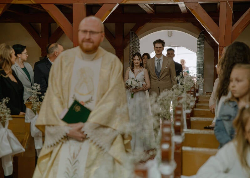 Fotografia ślubna Grudziądz – ślub kościelny Wiktorii i Dawida w Piaskach. Kolorowy kadr z ceremonii; ksiądz w złotym ornacie idzie przodem, a w tle widać parę młodą idącą nawą główną wśród gości weselnych.