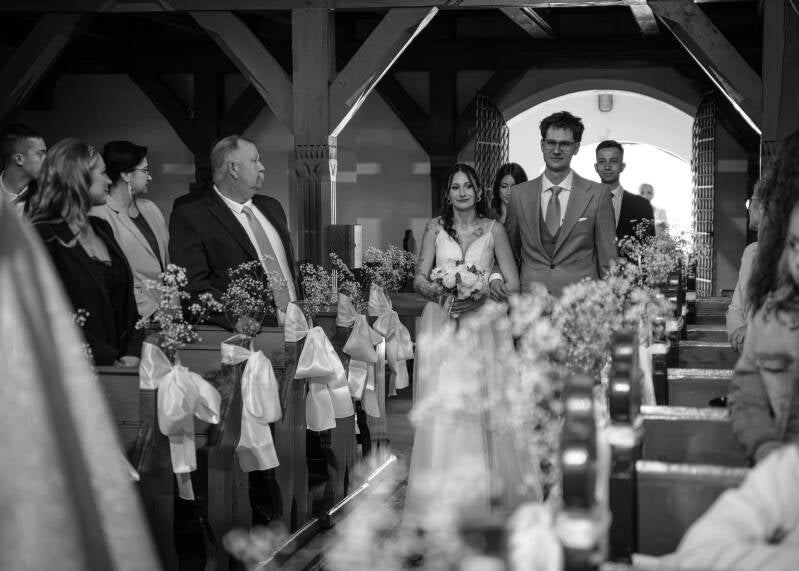Fotografia ślubna Grudziądz – reportaż z ceremonii ślubnej w kościele w Piaskach. Czarno-białe zdjęcie przedstawiające pannę młodą z bukietem i pana młodego idących nawą główną wśród uśmiechniętych gości weselnych