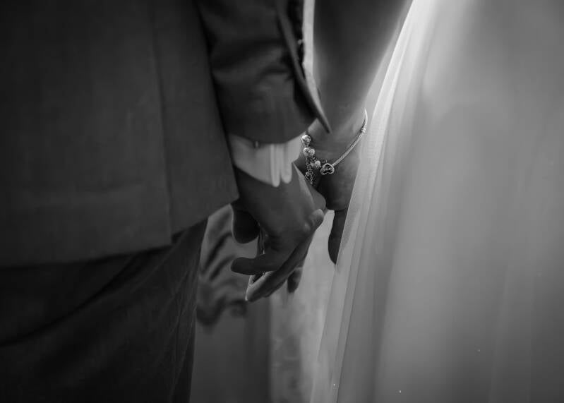 Fotografia ślubna Grudziądz – artystyczny detal ślubny z ceremonii w kościele w Piaskach. Czarno-białe makro ujęcie przedstawiające parę młodą trzymającą się mocno za ręce; widoczna elegancka bransoletka z zawieszkami na nadgarstku panny młodej i mankiet 