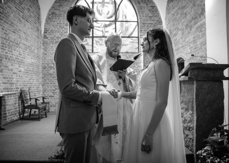 Fotografia ślubna Grudziądz – przysięga małżeńska Wiktorii i Dawida w kościele w Piaskach. Czarno-białe zdjęcie z ceremonii przedstawiające księdza wiążącego białą stułą dłonie pary młodej stojącej przed ołtarzem.
