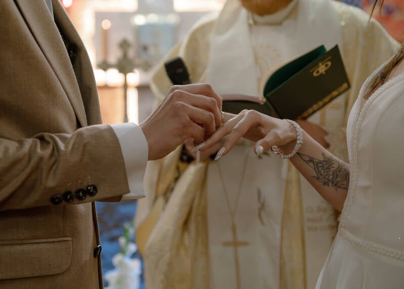 Fotografia ślubna Grudziądz – nałożenie obrączek ślubnych przez Wiktorię i Dawida w kościele w Piaskach. Bliski kadr w ciepłych barwach przedstawia pana młodego wsuwającego złotą obrączkę na palec panny młodej, ubranej w suknię z perełkami, z widocznym ta