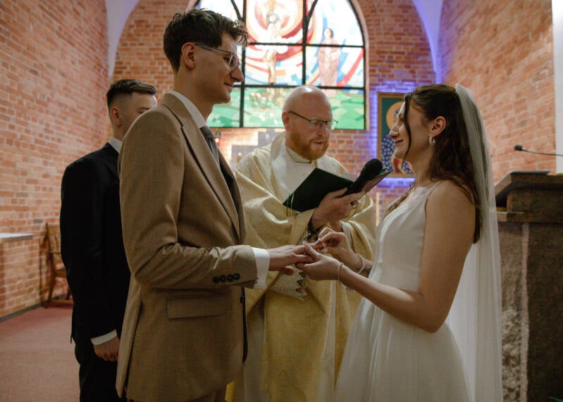 Fotografia ślubna Grudziądz – wzajemna wymiana obrączek przez Wiktorię i Dawida podczas ślubu w kościele w Piaskach. Kolorowy kadr przedstawia pannę młodą wsuwającą obrączkę na palec pana młodego przed ołtarzem, w obecności księdza i świadka.