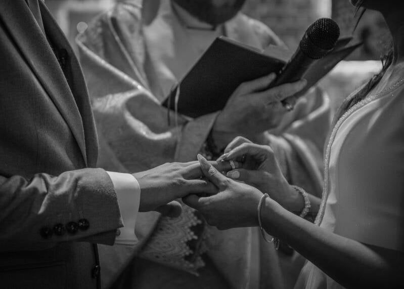 Fotograf ślubny Grudziądz i okolice – detaliczny reportaż ślubny z ceremonii blisko Torunia i Bydgoszczy. Zbliżenie na dłonie nowożeńców podczas sakramentalnej wymiany obrączek przed ołtarzem, w tle widoczny ksiądz z liturgiczną księgą.